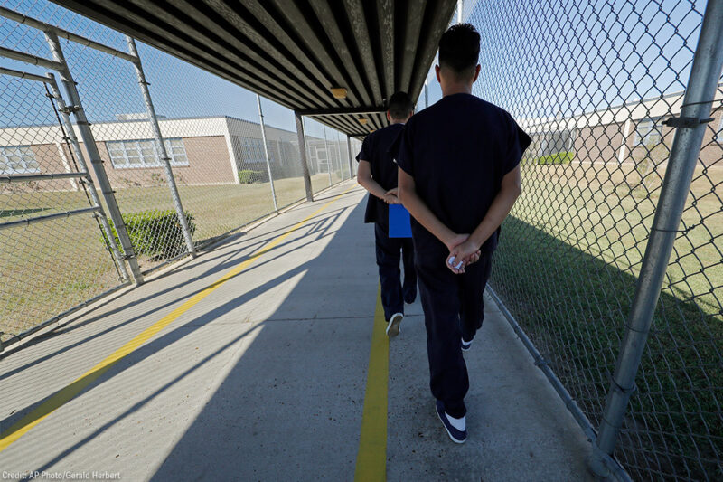 Immigrant detainees walk with their hands clasped behind their backs along a line painted on a walkway