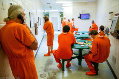 Inmates pass the time in their cell block. Some pictured on the phone, some watching television, and some playing card games at tables. As concerns regarding the spread of COVID-19 mount, inmates argue that prisons have no plan in place protect against the spread of the illness.