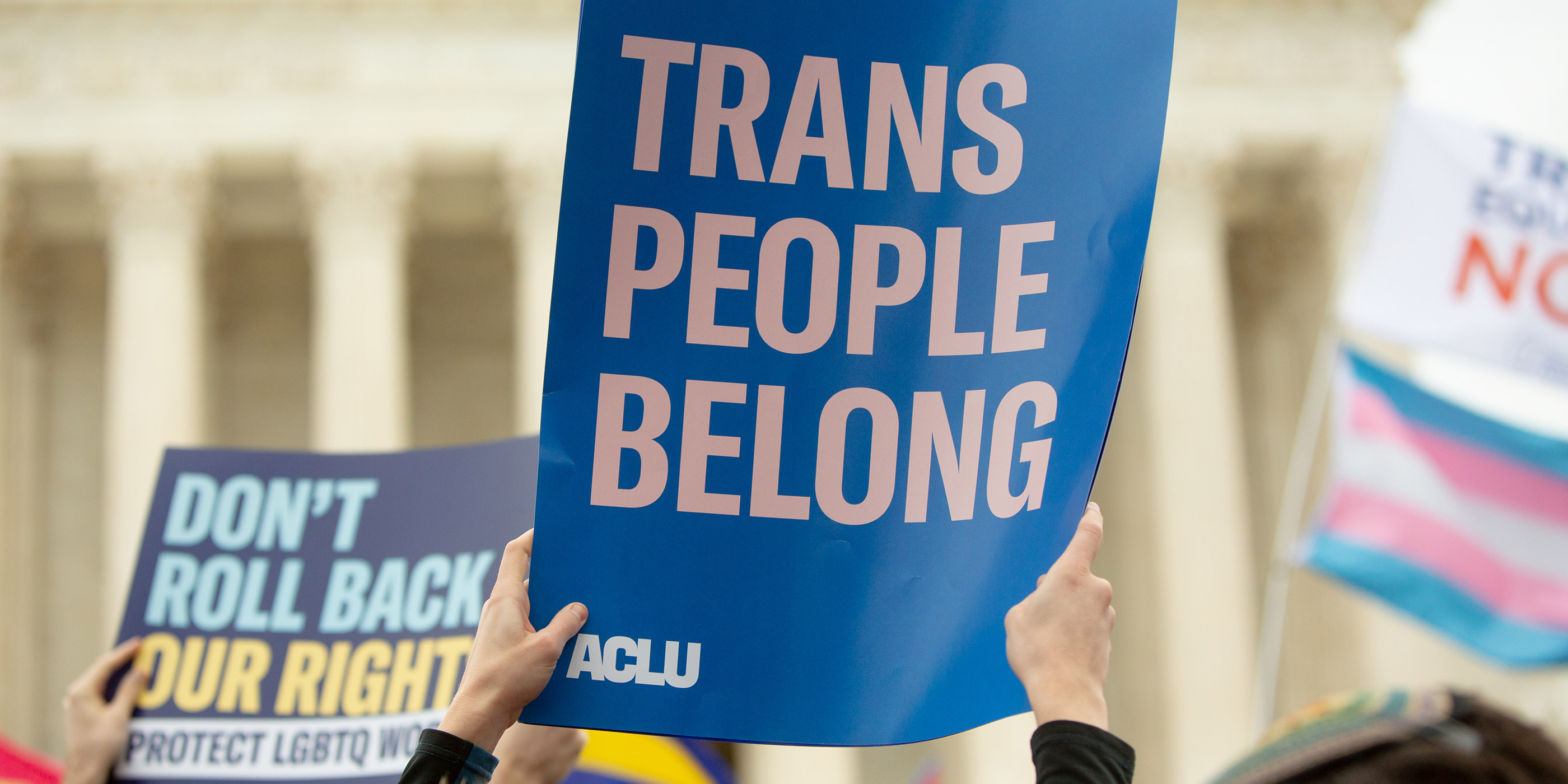 Someone holding up a poster in front of the Supreme Court that reads "trans people belong" and in the background, another person holds a poster that says "don't roll back our rights." This is in the face of trans discrimination and at a time when SCOTUS has a number of cases on the docket that affect trans lives.