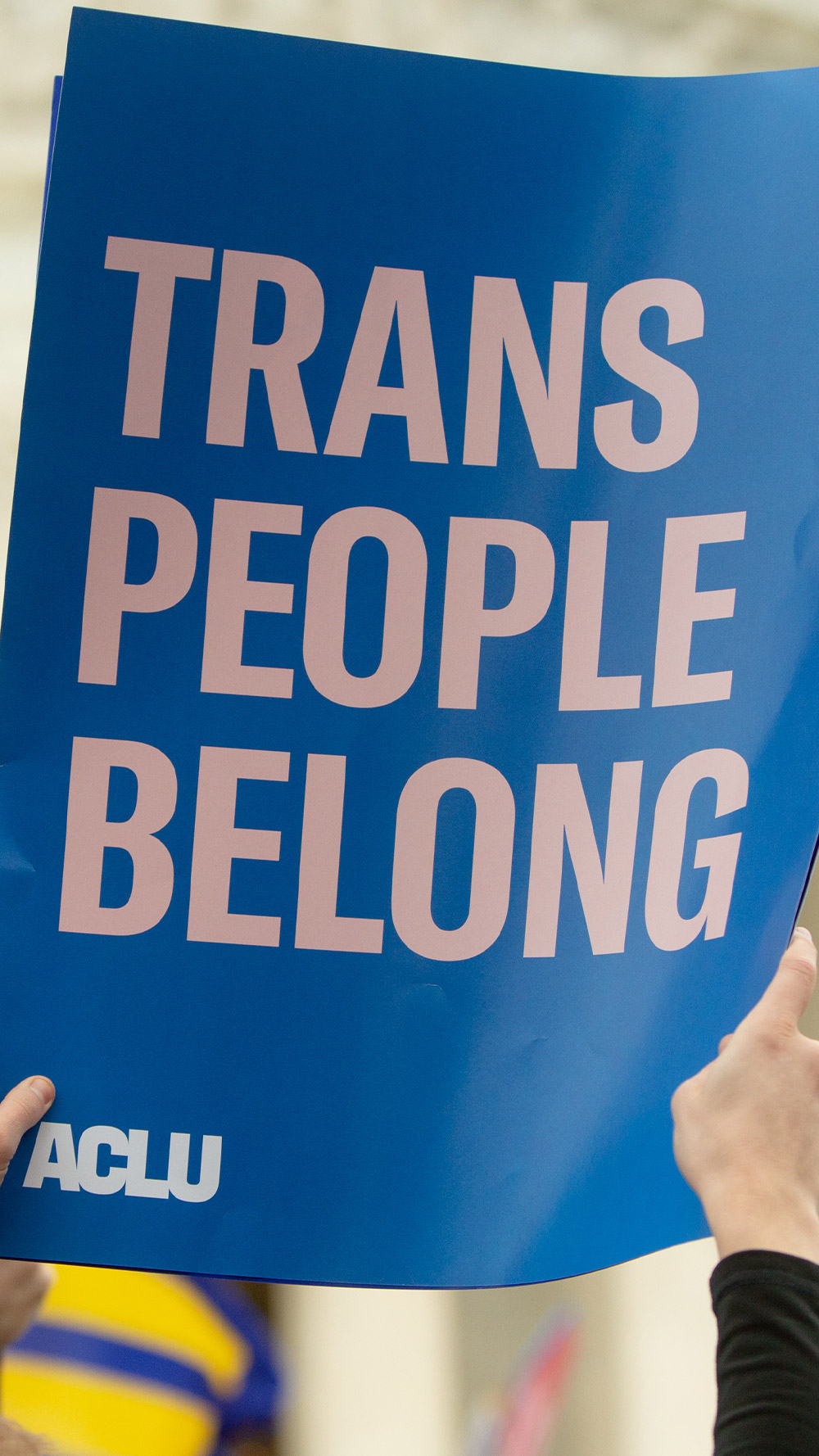 Someone holding up a poster in front of the Supreme Court that reads "trans people belong" and in the background, another person holds a poster that says "don't roll back our rights." This is in the face of trans discrimination and at a time when SCOTUS has a number of cases on the docket that affect trans lives.