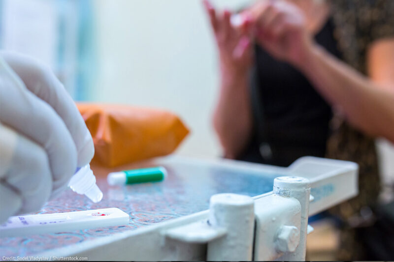 A gloved hand adds reagent to a COVID-19 serological antibody test with the patient sitting nearby.