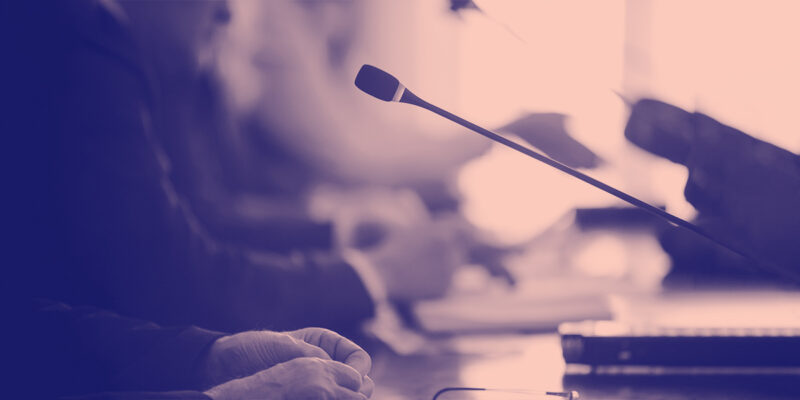 Pink tinted image of a lawmaker's hands placed on a desk in front of a mic.