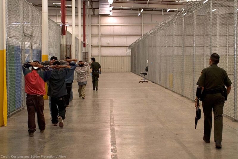 Immigrant detainees walk down a hall escorted by heavily armed CBP officers.