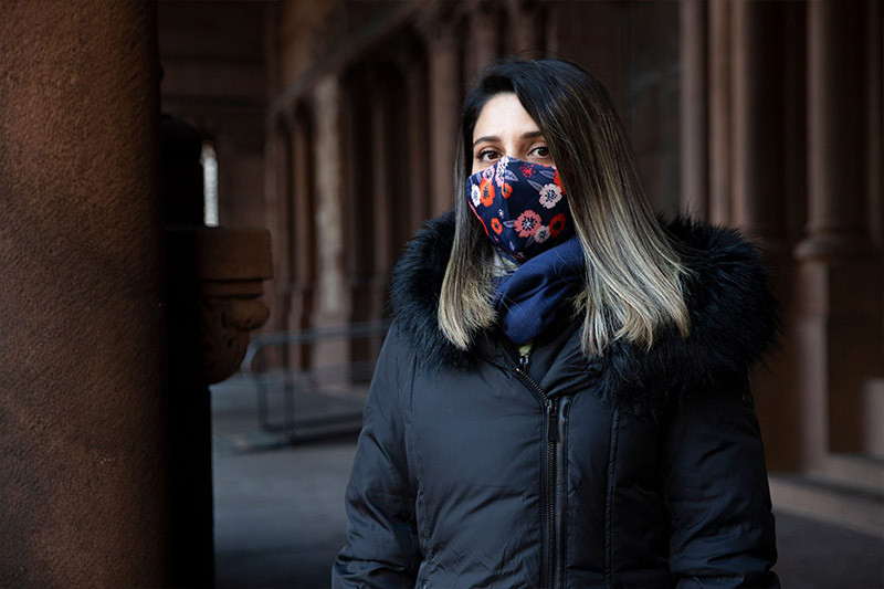 Woman in black coat and mask standing in front of columns