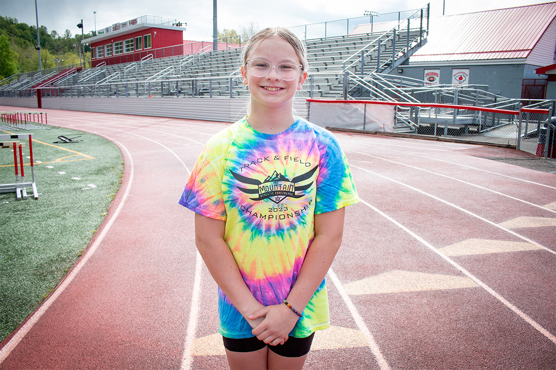 Photo of Becky, a white girl with blonde hair wearing clear-framed glasses and a tie-dyed rainbow t-shirt.