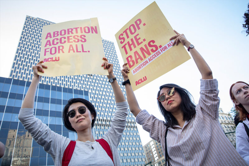 Two women holding up signs that say, "Abortion access for all." and "Stop. The. Bans." respectively.