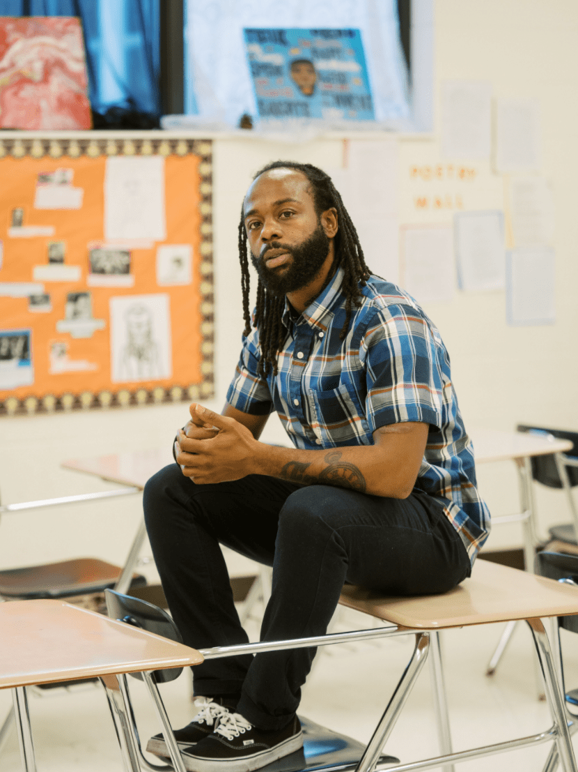 Anthony, teacher, is sitting on top of an empty student desk