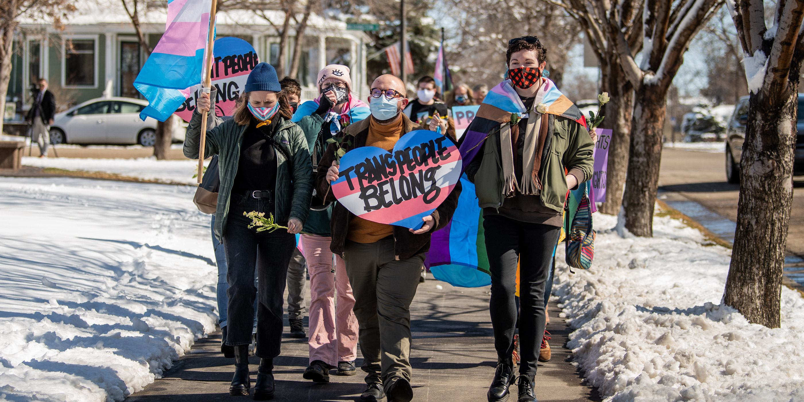 Individuals at a rally march, with a sign that reads “Trans People Belong.”