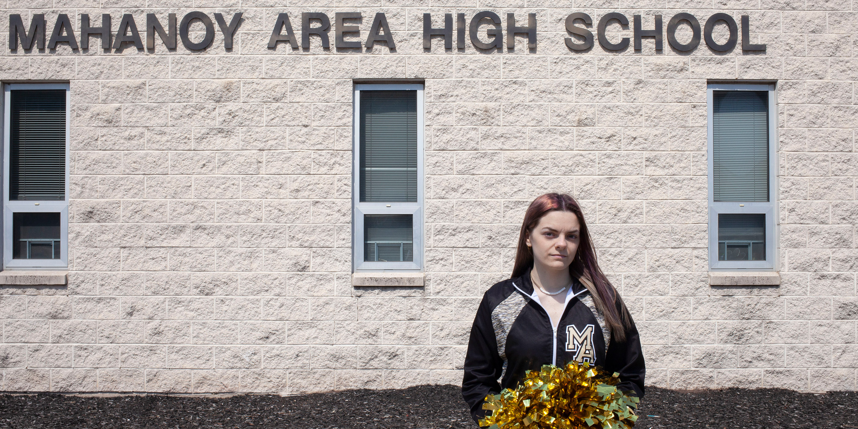 A photo of Brandi Levy holding gold pom-poms in front of Mahanoy Area High School.