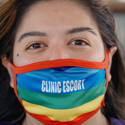 A close-up of Mariceli Alegria who's wearing a rainbow facemask with the printed words "Clinic Escort".