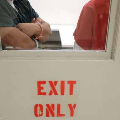 Two detainees seen waiting through a window behind closed doors.