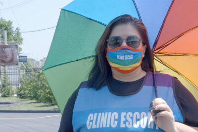Abortion clinic escort Mariceli Alegria under the shade of an umbrella and wearing a rainbow facemask with the printed words