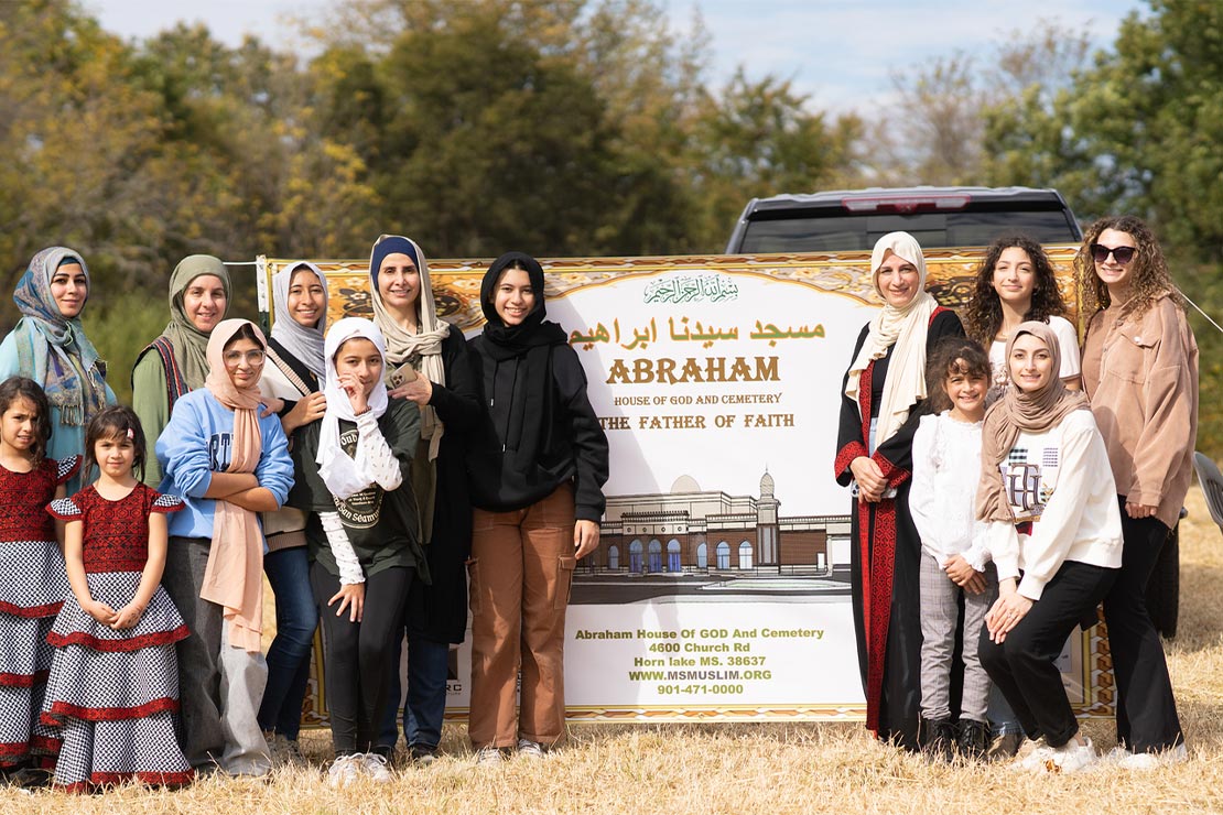 Female members of the Abraham House of God (the first mosque in DeSoto County Mississippi) of all ages pose for a picture, standing on the left and right of a sign saying "Abraham House pof God and Cemetery - The Father of Faith".