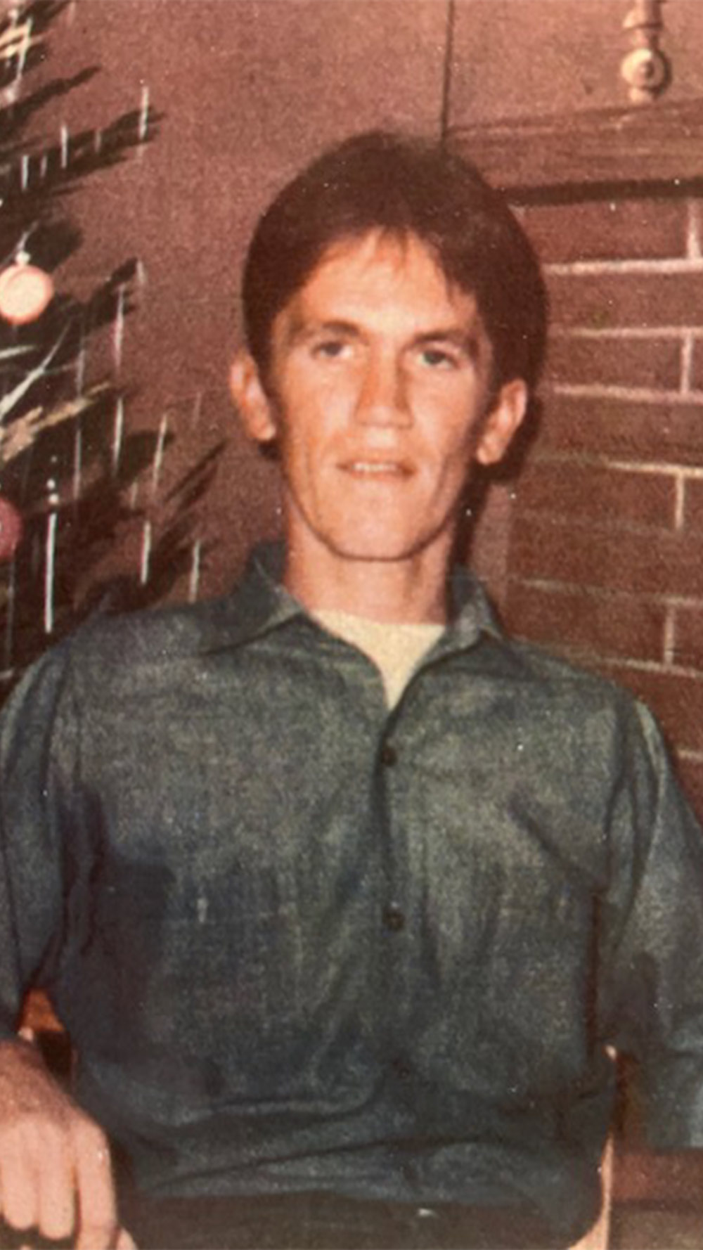 In front of a painted background of a painted Christmas tree on the left and fireplace on the right, Shawn Jensen sits for the camera as he spends his first Christmas in Arizona State Prison.