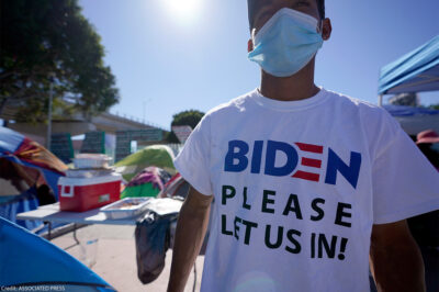 A man seeking asylum in the United States wears a shirt that reads, "BIDEN PLEASE LET US IN!," as he stands among tents that line an entrance to the border crossing in Tijuana, Mexico.