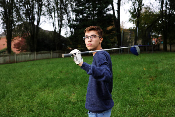 Photo of Luc Esquivel, a 14-year-old boy in a blue sweater and glasses with a golf club over his shoulder. Luc is standing in a green yard.