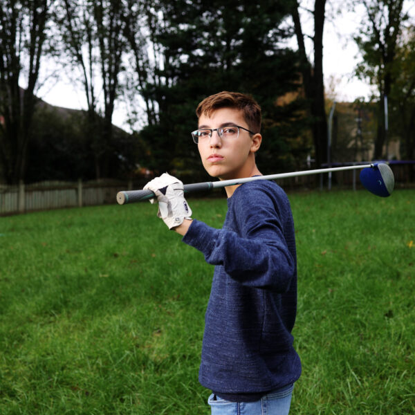 Photo of Luc Esquivel, a 14-year-old boy in a blue sweater and glasses with a golf club over his shoulder. Luc is standing in a green yard.