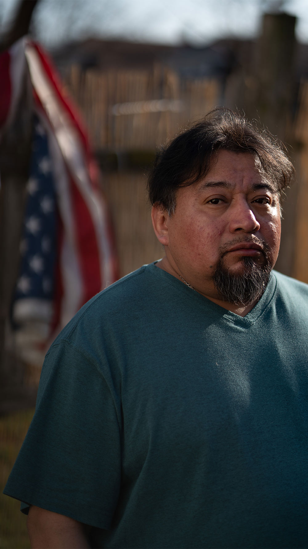 Margarito Castañon Nava looks into the camera as an American flag hangs from a wooden fence behind him.
