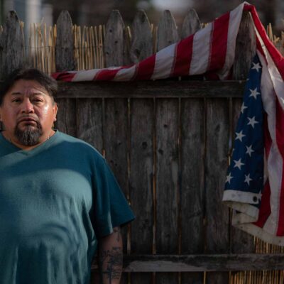 Margarito Casta​​ñon Nava looks into the camera as he stands next to an American flag hanging from a wooden fence behind him on his right.