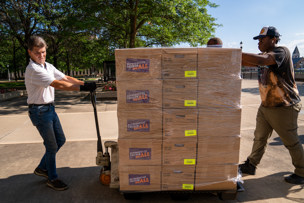 Two men wheeling a large stack of boxes filled with signatures.