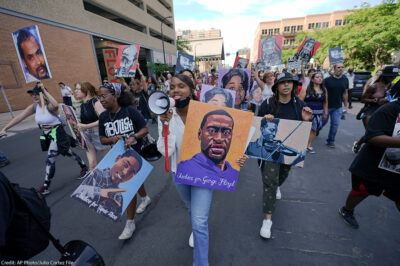 Protestors walking down the street, with one holding a microphone and a sign with art of George Floyd's face.