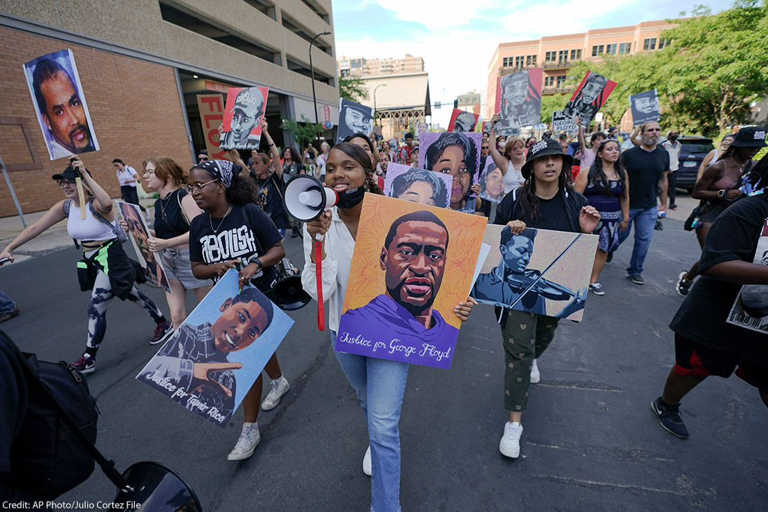 Protestors walking down the street, with one holding a microphone and a sign with art of George Floyd's face.