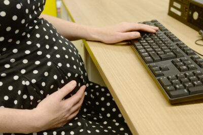 Pregnant person sits at a desk with one hand resting on their stomach and the other on a keyboard.