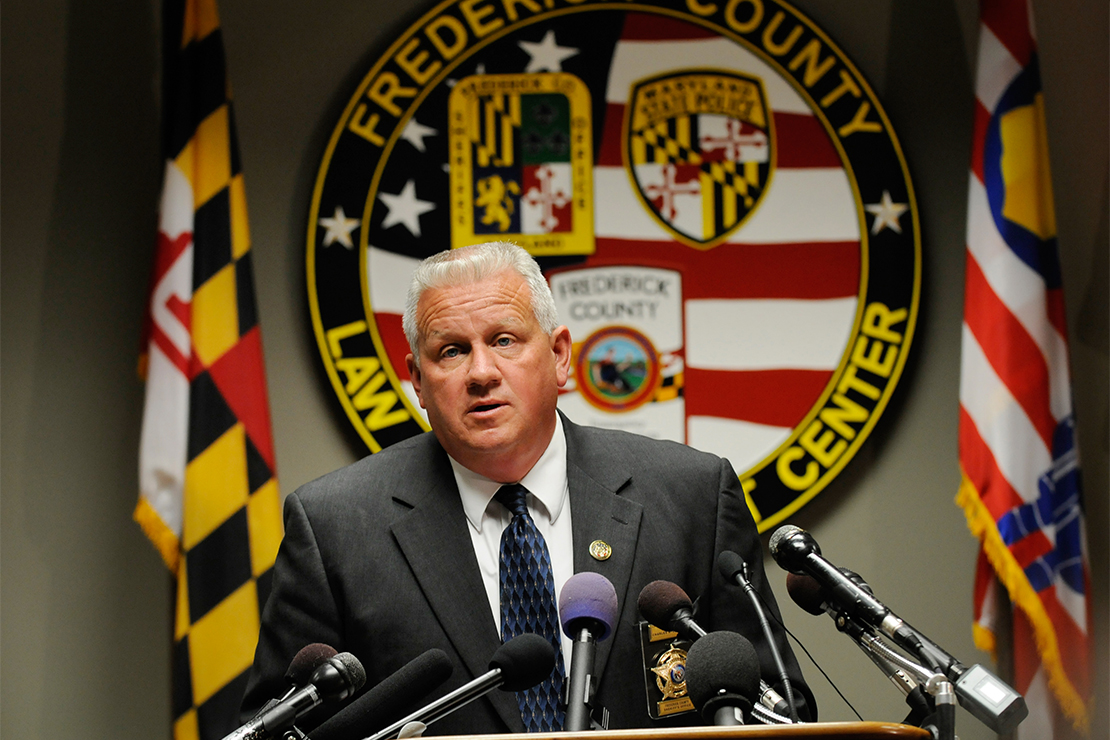 Frederick County Sheriff Chuck Jenkins standing in front of a podium filled with microphones with a giant Frederick County seal behind him.