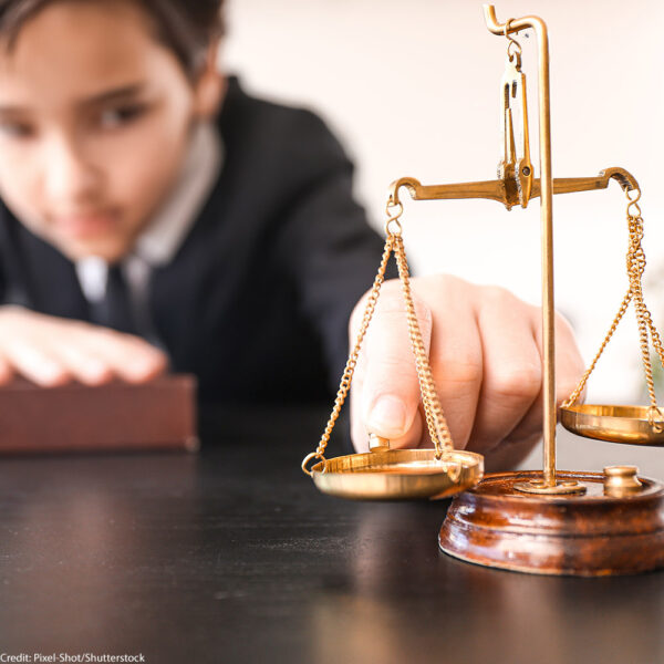 A child dressed in a suit with a hand on top of a book, tips scales of justice.