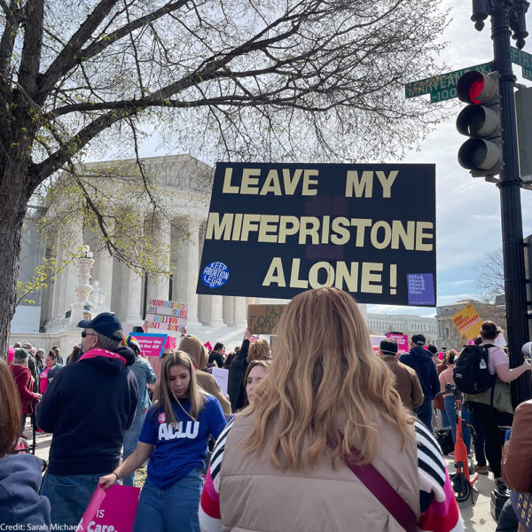 a sign saying "leave my mifepristone alone!" is foregrounded with protesters in front of the supreme court.