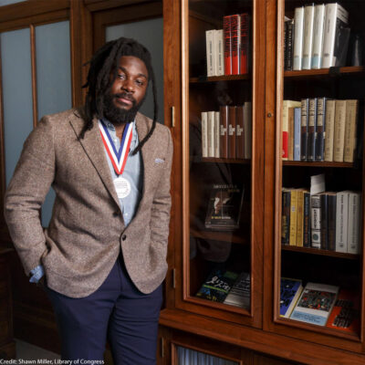 Jason Reynolds, National Ambassador for Young People’s Literature, 2020-2022. Stands in front of a bookshelf.